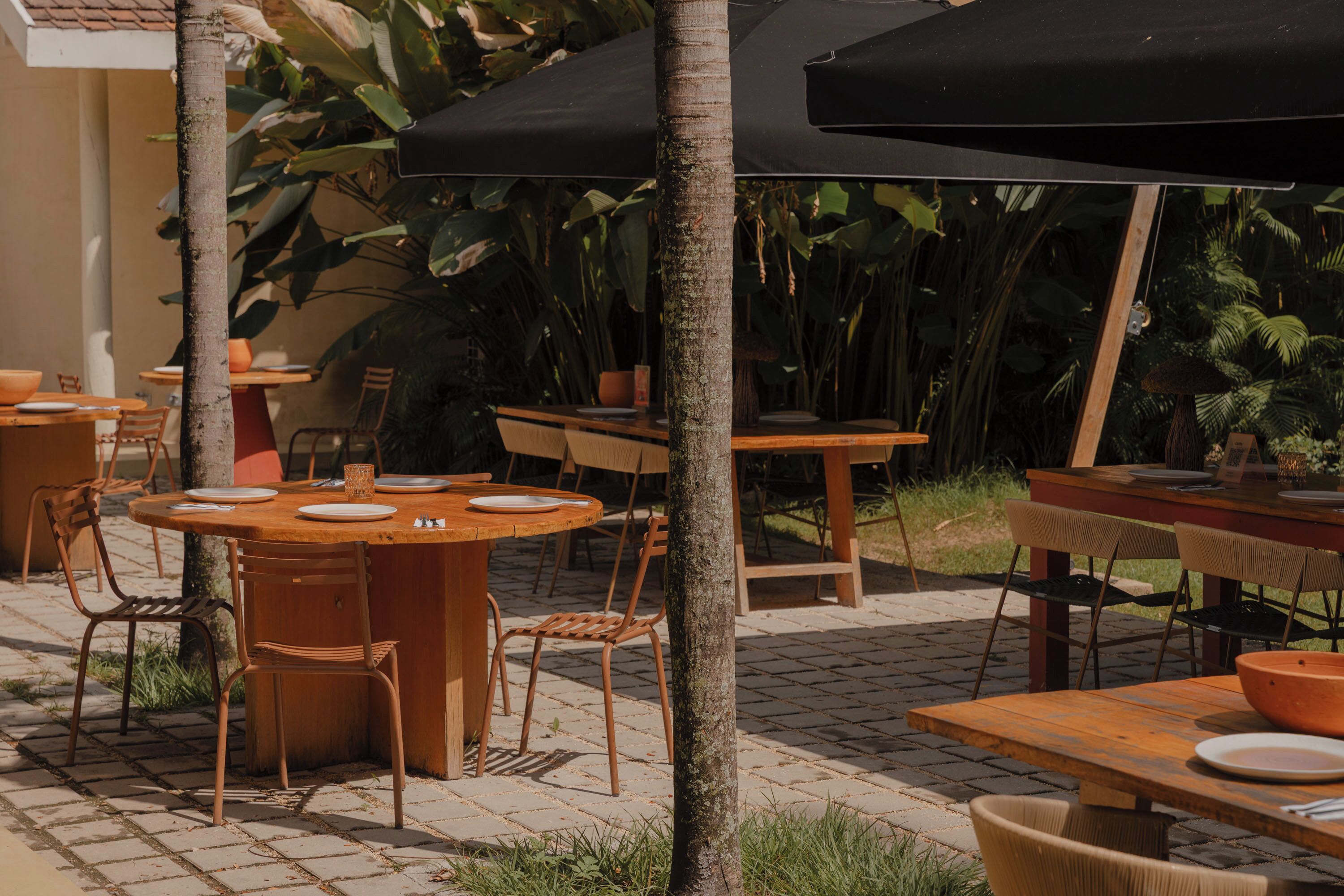 Un área de comedor al aire libre con mesas y sillas de madera, rodeada de plantas tropicales y un piso de baldosas.