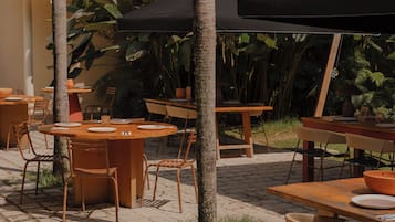 Un área de comedor al aire libre con mesas y sillas de madera, rodeada de plantas tropicales y un piso de baldosas.