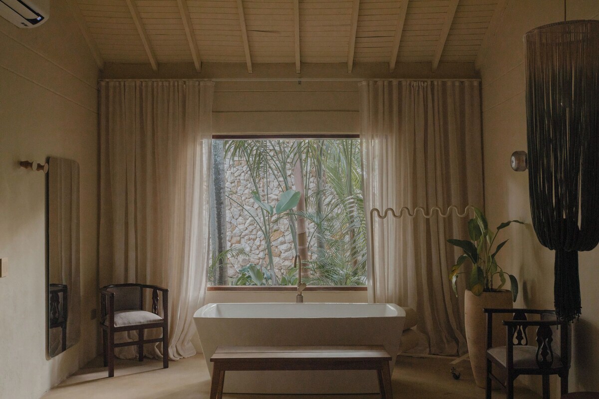 A bathroom with a bathtub, a window with curtains, a chair, and a potted plant.