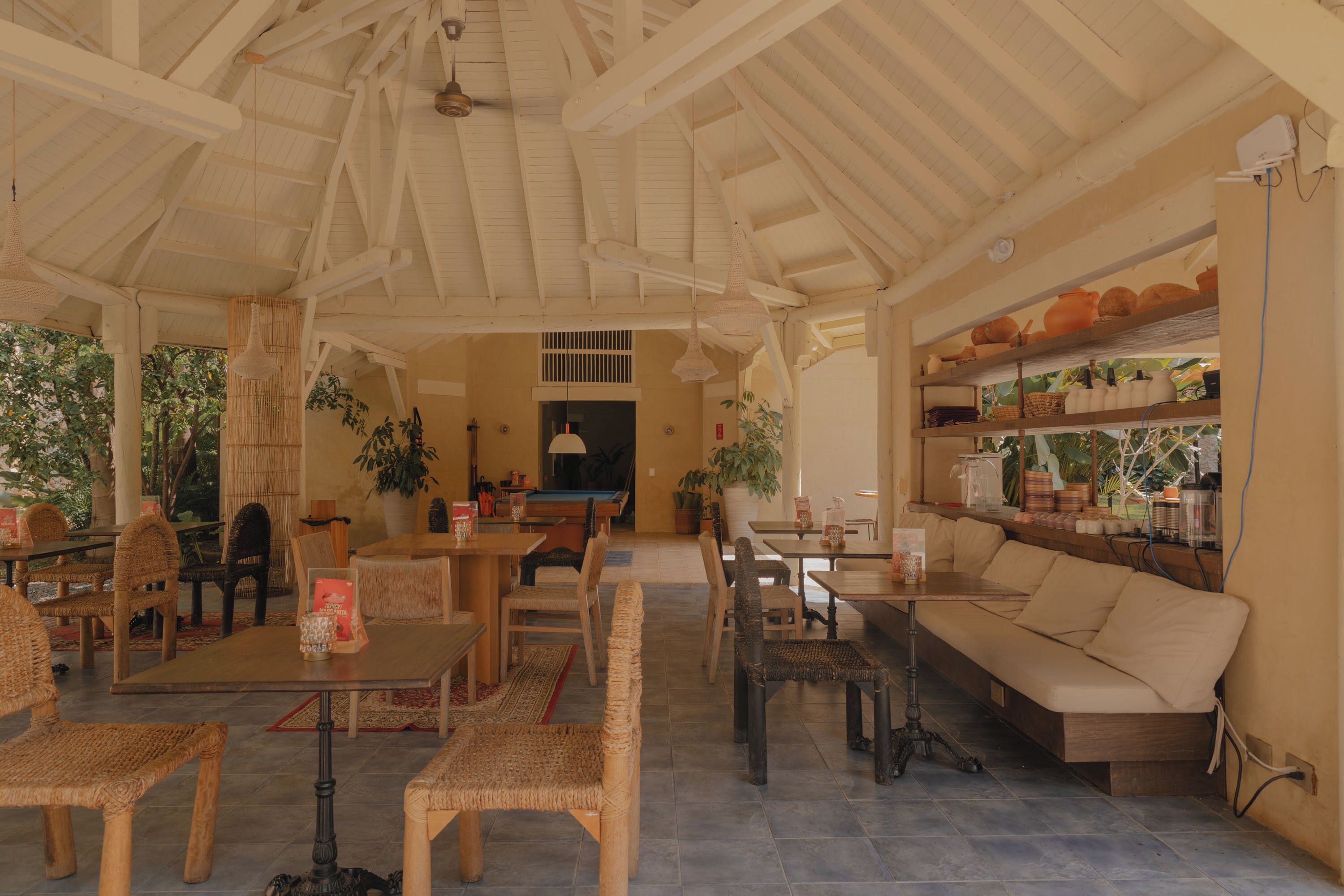 A spacious indoor area with a high ceiling, wooden beams, and a pool table. There are several seating areas with wicker chairs and tables, and a sofa along the wall.