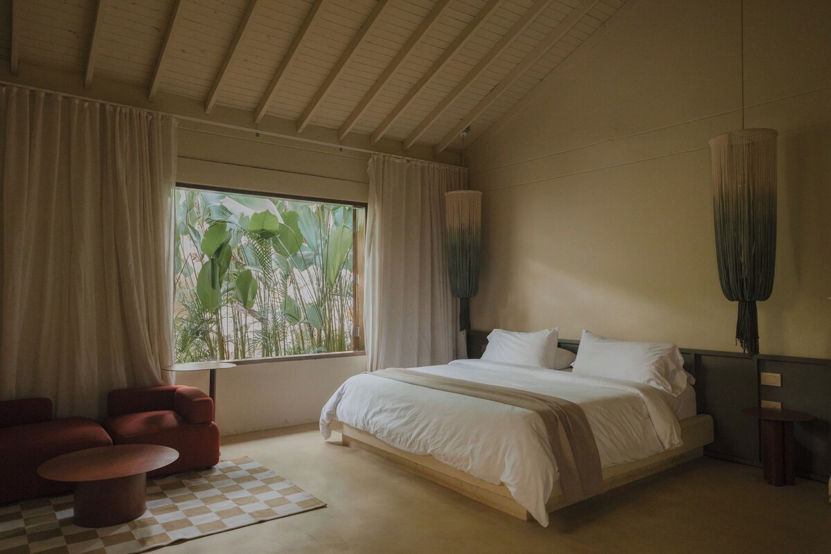 A bedroom with a large bed, a chair, a checkered rug, and a window with curtains.