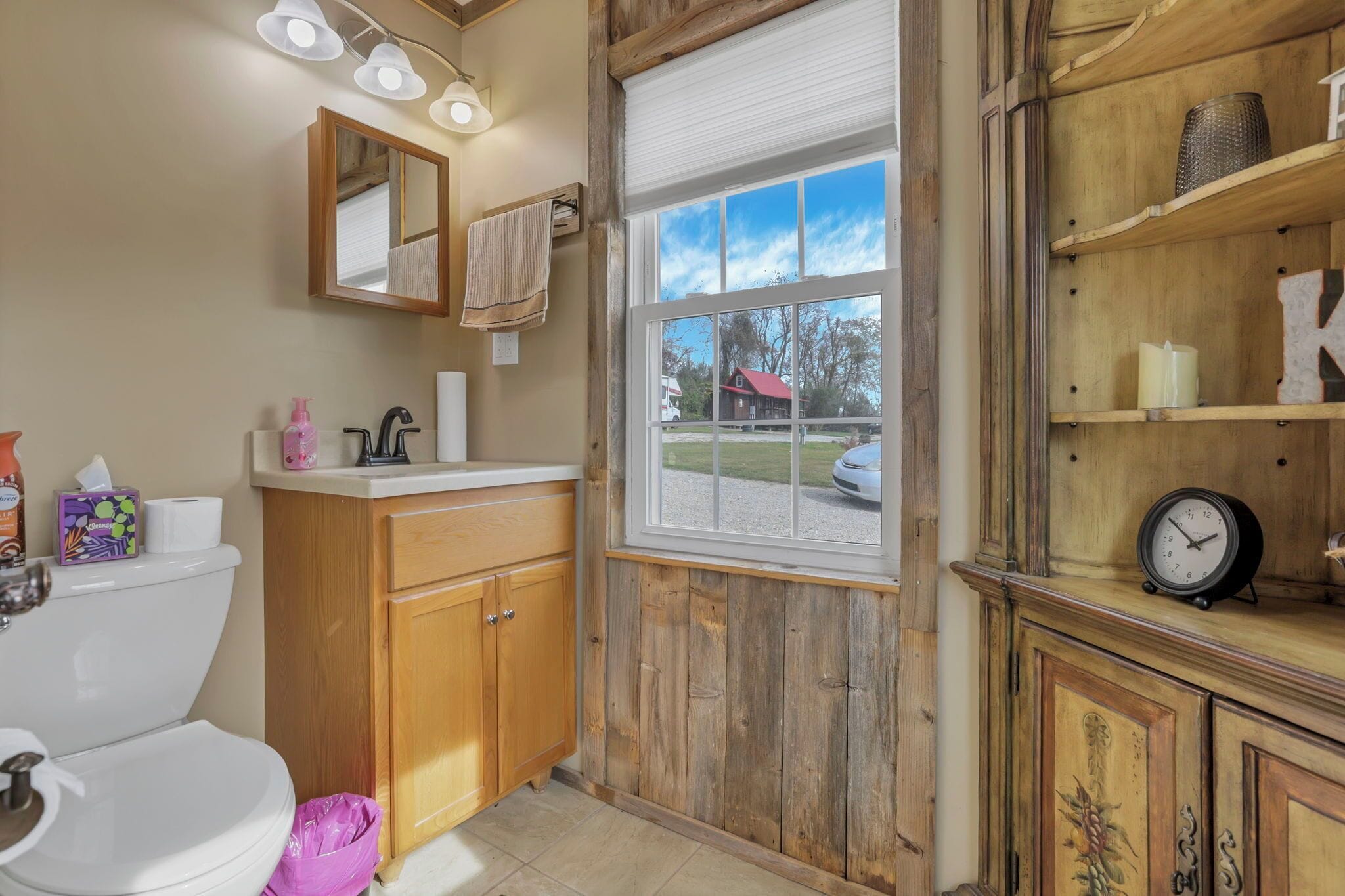A bathroom with wooden cabinets, a toilet, a sink, a mirror, and a window with a view of a house.