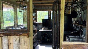A rustic wooden cabin interior with a kitchen area, a large window, and a view of greenery outside. - Cabin with Character (New Lexington)