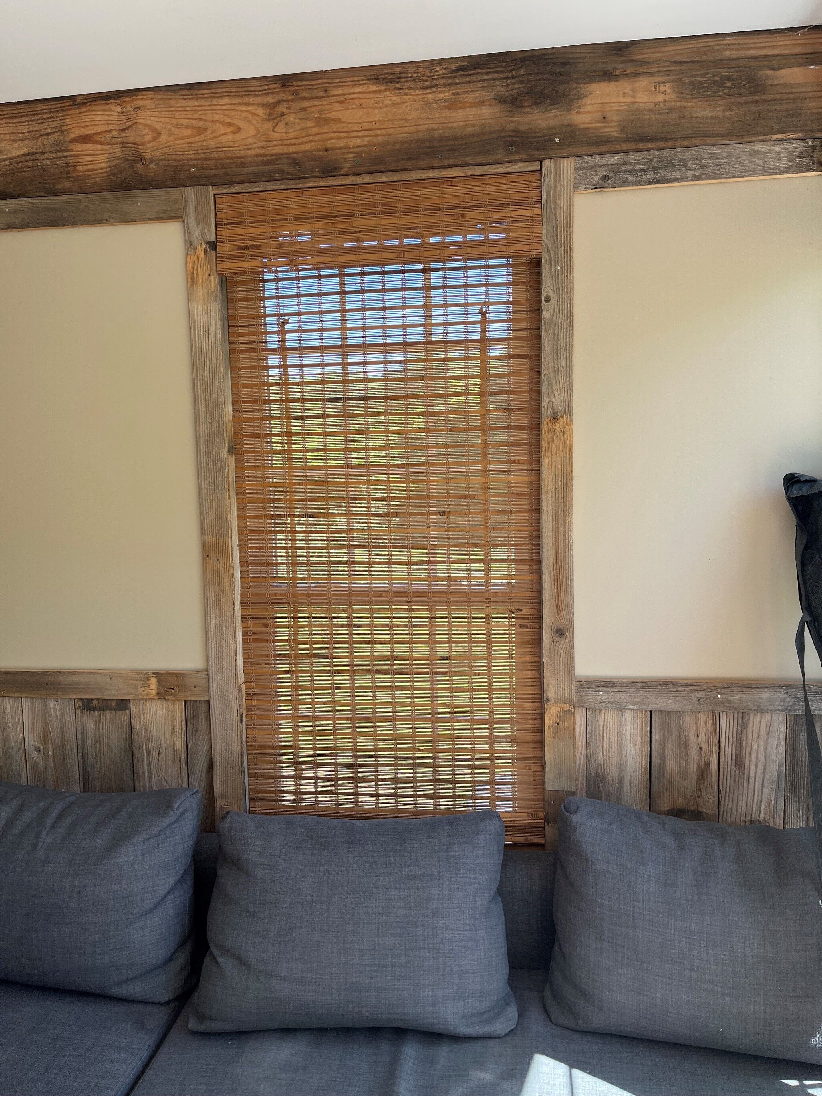 A room with a wooden ceiling, a window with horizontal blinds, and a grey sofa with cushions.