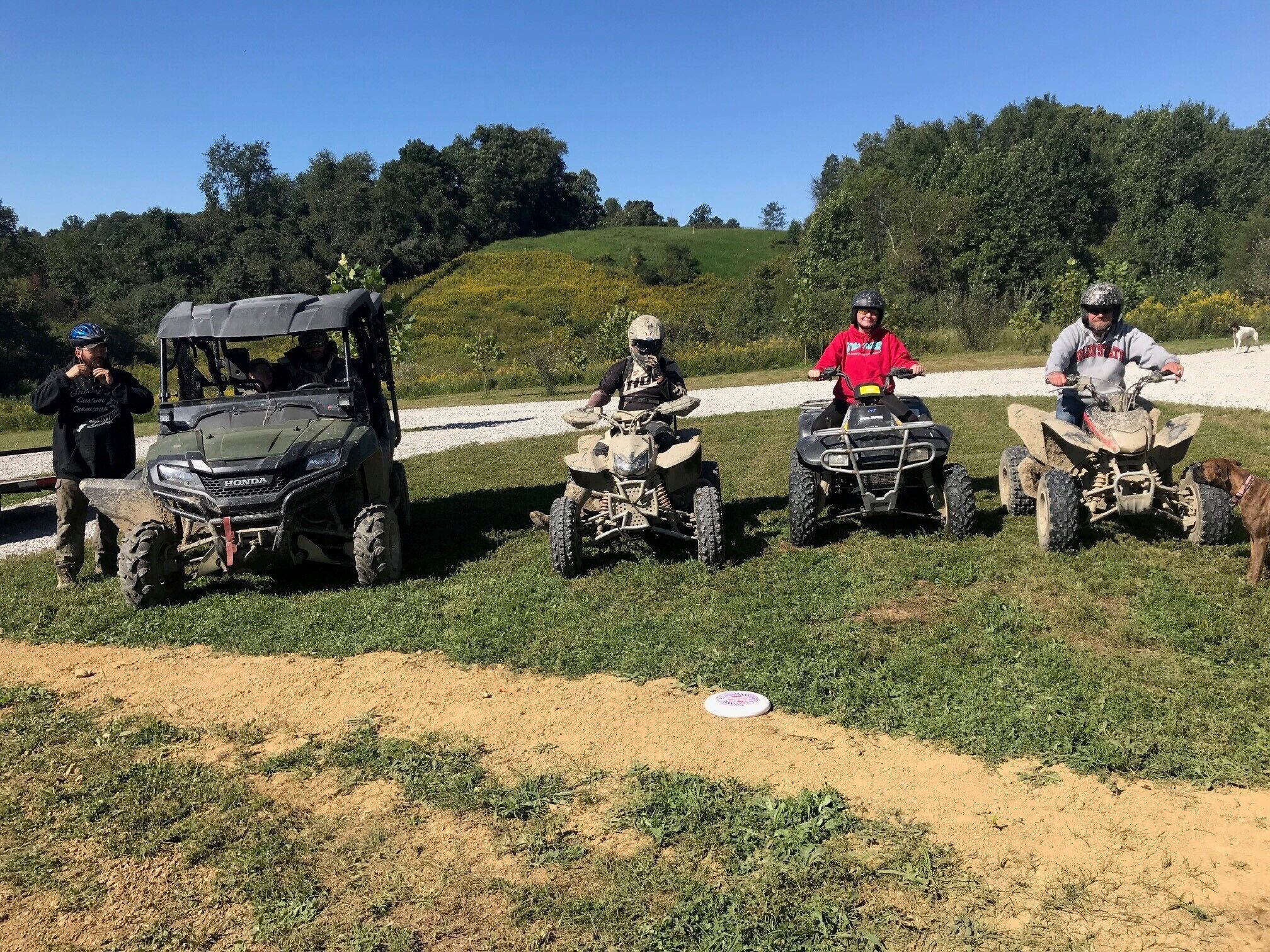 A group of people on ATVs in a grassy area with a dirt path and trees in the background.