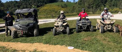Un groupe de personnes conduisant des quads dans une zone herbeuse, avec un chemin de terre et des arbres en arrière-plan.