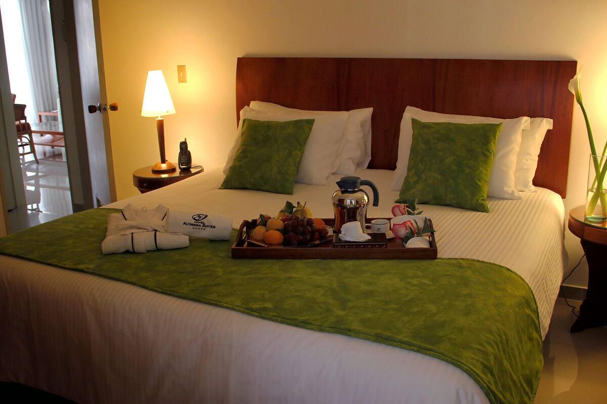 Een hotelkamer met een netjes opgemaakt bed, een schaal met fruit en een koffiezetapparaat, en een nachtkastje met een lamp en een bloemenvaas.