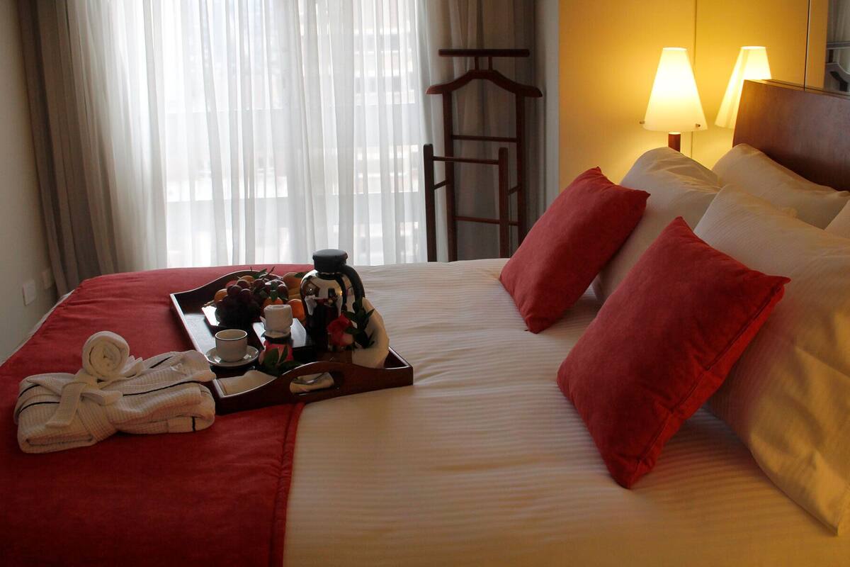 Een netjes opgemaakt bed met rode en witte kussens, een dienblad met fruit en een fles, en een handdoek ernaast.