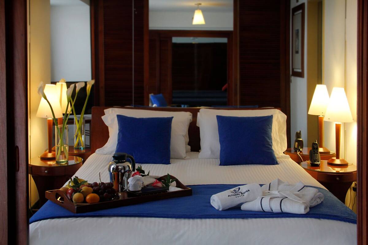 Een hotelkamer met een groot bed, twee blauwe kussens, een dienblad met fruit en een koffiezetapparaat, en twee bedlampjes.