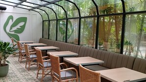 Un restaurant à l’architecture inspirée d’une serre, avec des tables et des chaises en osier, et un grand logo vert sur le mur.