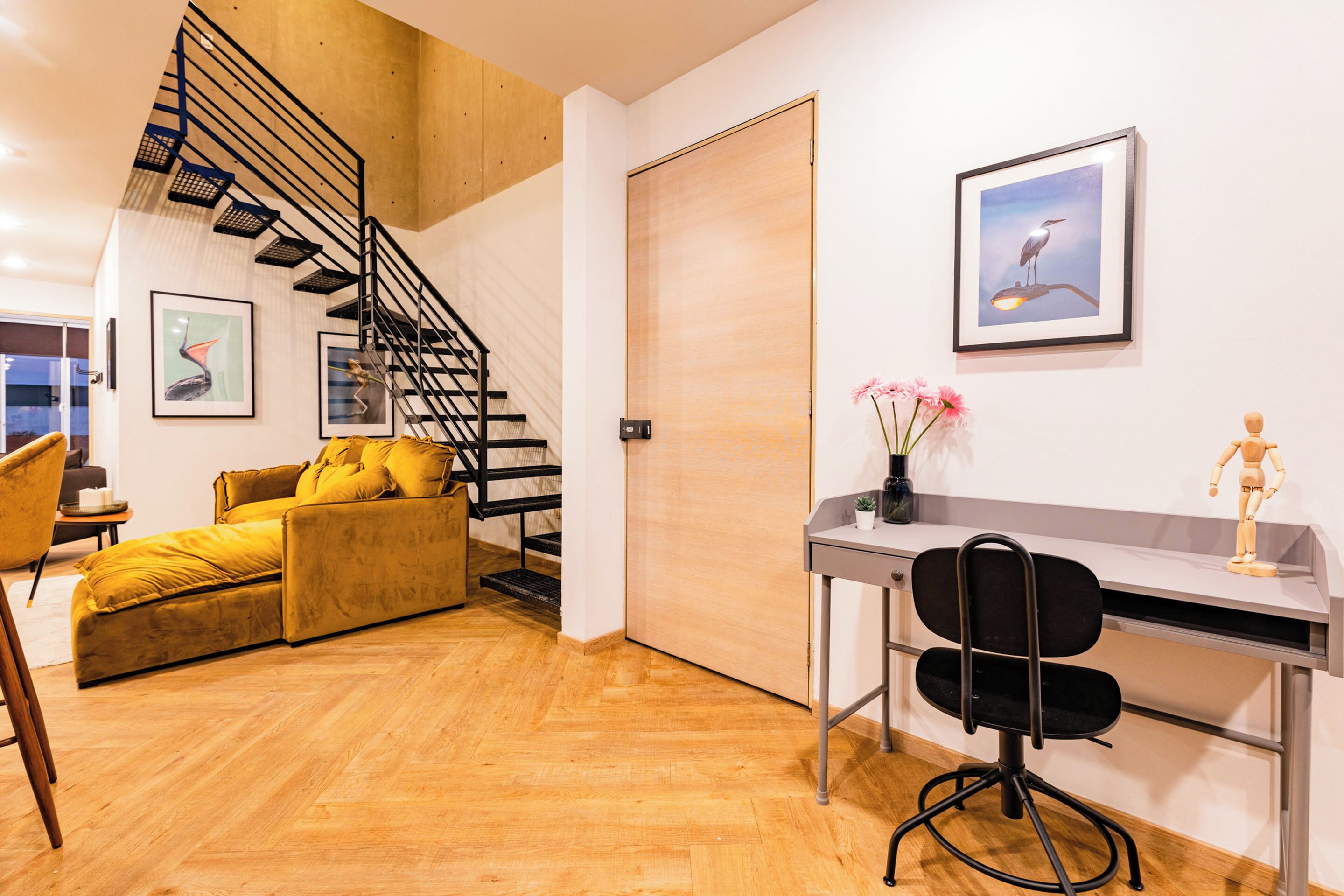 A modern living room with a wooden floor, a yellow sofa, a black office chair, a desk, and a staircase.