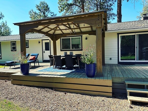 Une terrasse extérieure couverte, dotée d’un sol en bois, d’une table avec des chaises et de plantes en pot.