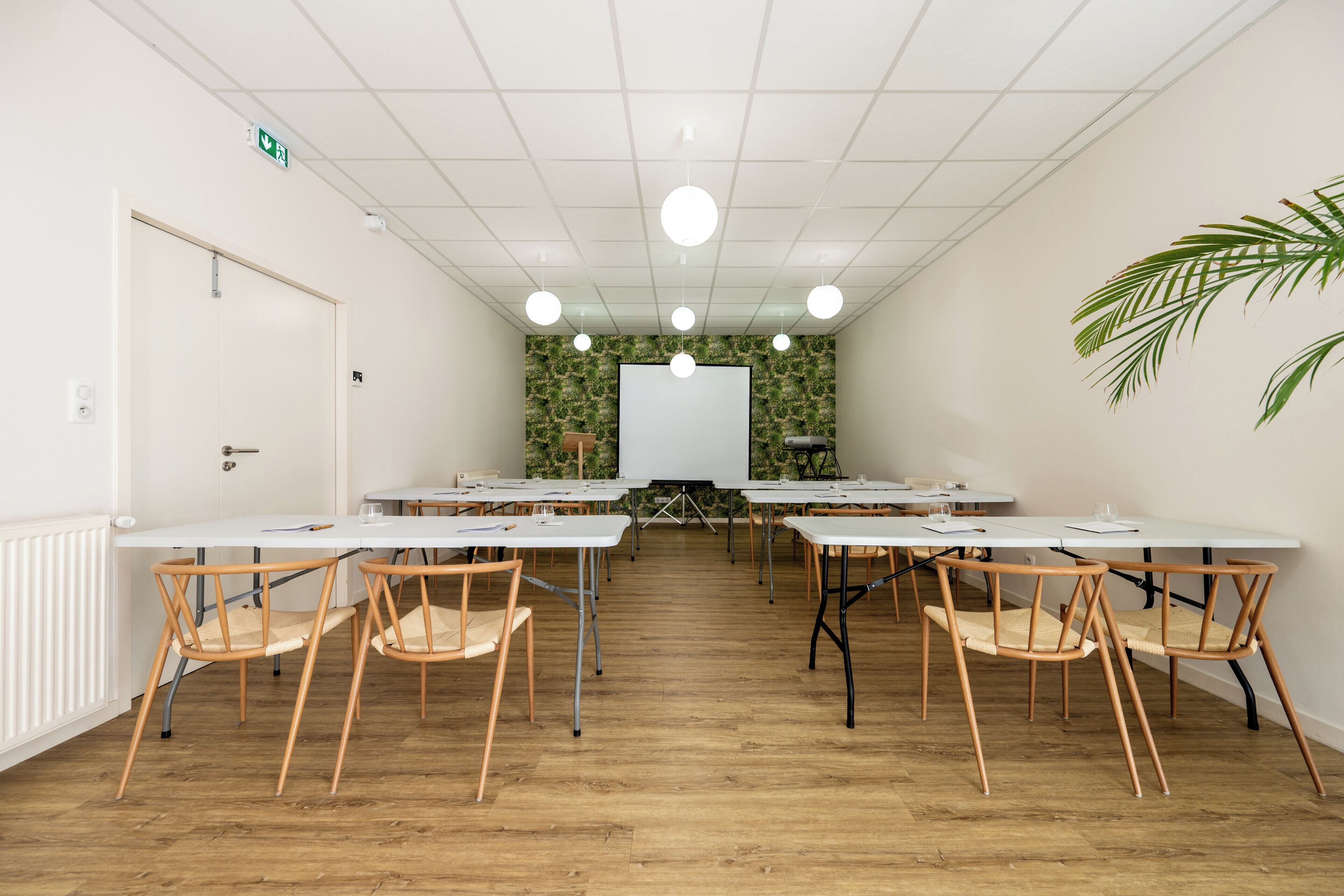 Uma sala de conferências com mesas brancas, cadeiras de madeira, um quadro branco e um painel decorativo com folhagens verdes.
