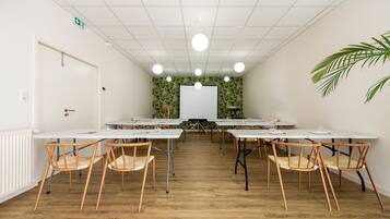 Uma sala de conferências com mesas brancas, cadeiras de madeira, um quadro branco e um painel decorativo com folhagens verdes.
