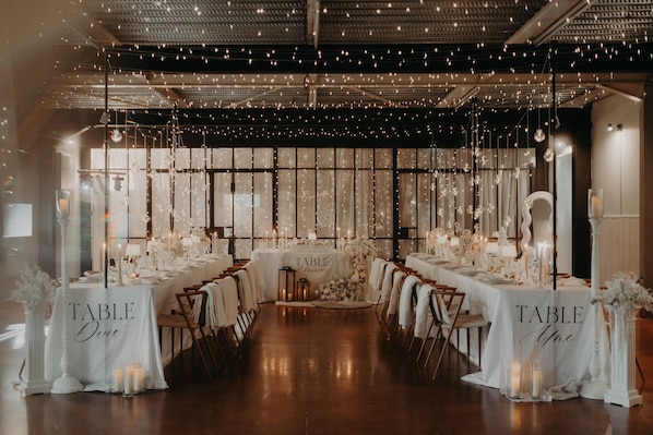 A long table with 'TABLE One' signage, surrounded by chairs and candles, in a room with a high ceiling and string lights.