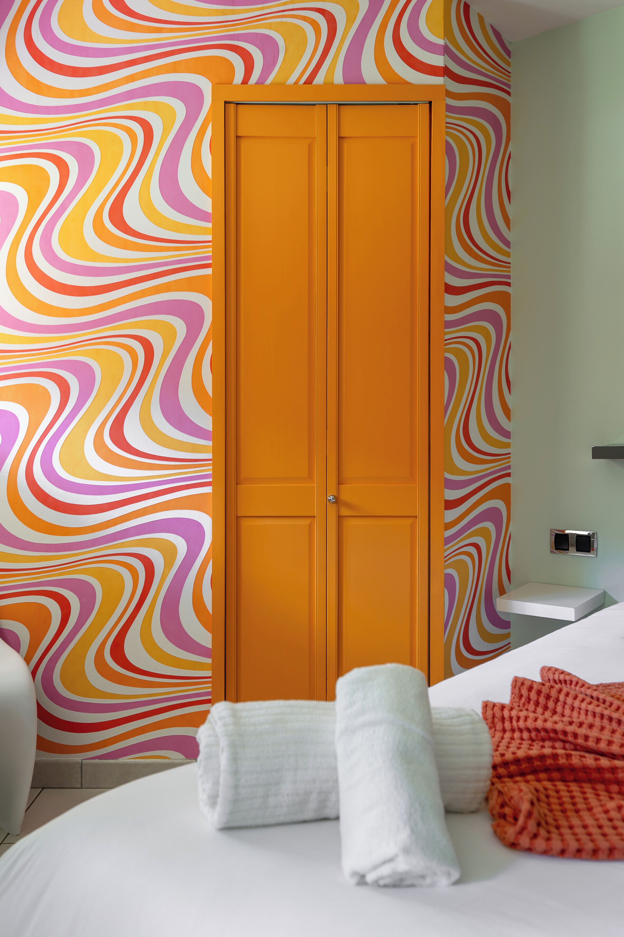 Une chambre d’hôtel avec un mur aux motifs colorés, une porte à deux battants, un lit recouvert de draps blancs et une petite table de chevet.