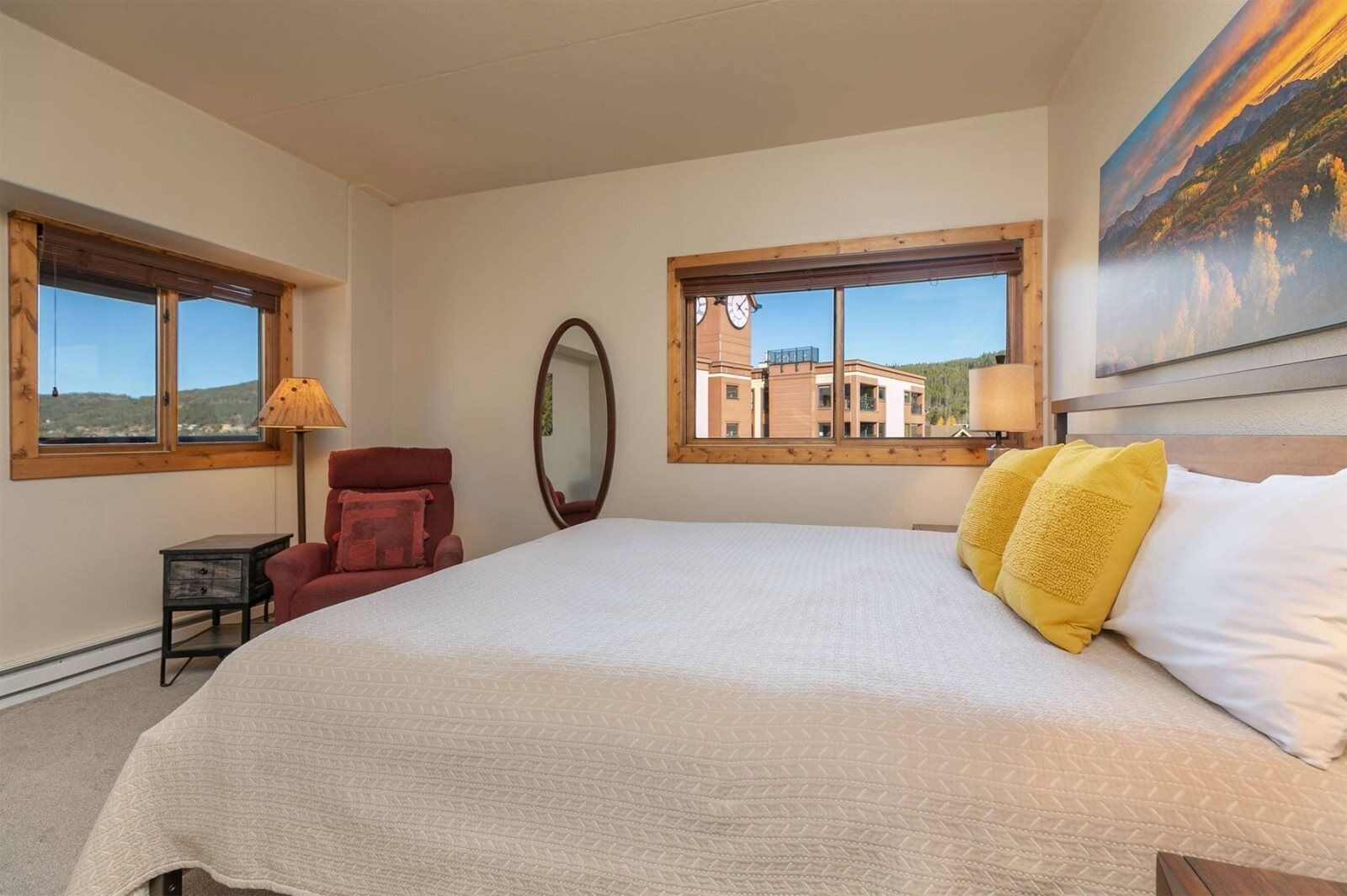 A bedroom with a large bed, two windows, a chair, a small table, and a painting on the wall.