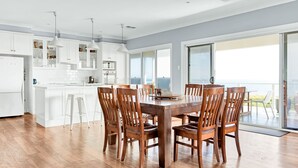 A modern kitchen with white cabinets, a dining area with wooden chairs, and a large sliding glass door leading to a balcony with a view of the ocean.