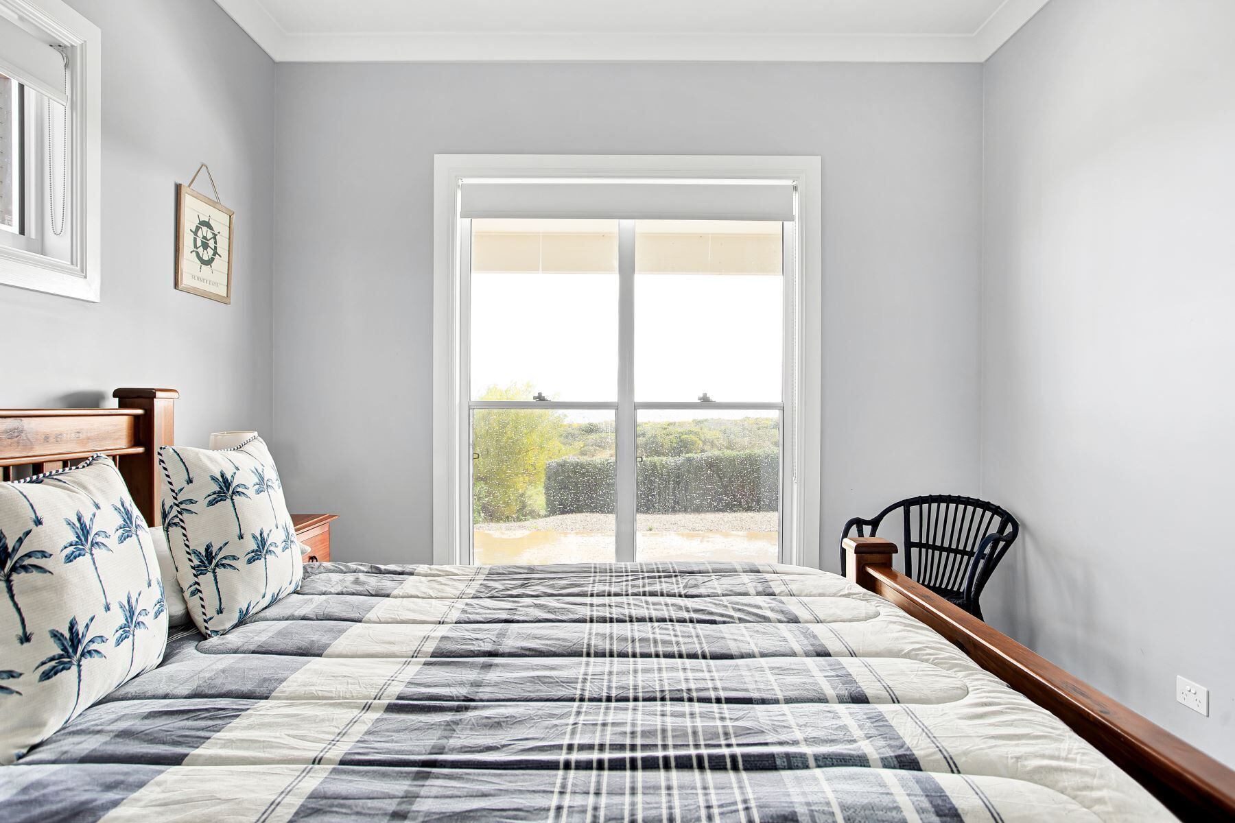 A bedroom with a large bed, a window with a view of greenery, a black chair, and a framed picture on the wall.