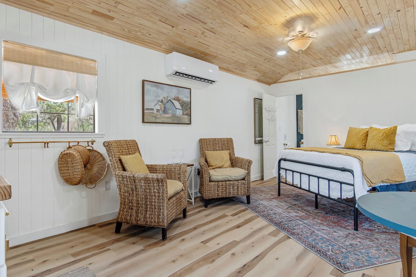 A room with a bed, two wicker chairs, a window with curtains, a painting, and a ceiling fan.