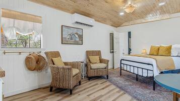 A room with a bed, two wicker chairs, a window with curtains, a painting, and a ceiling fan.