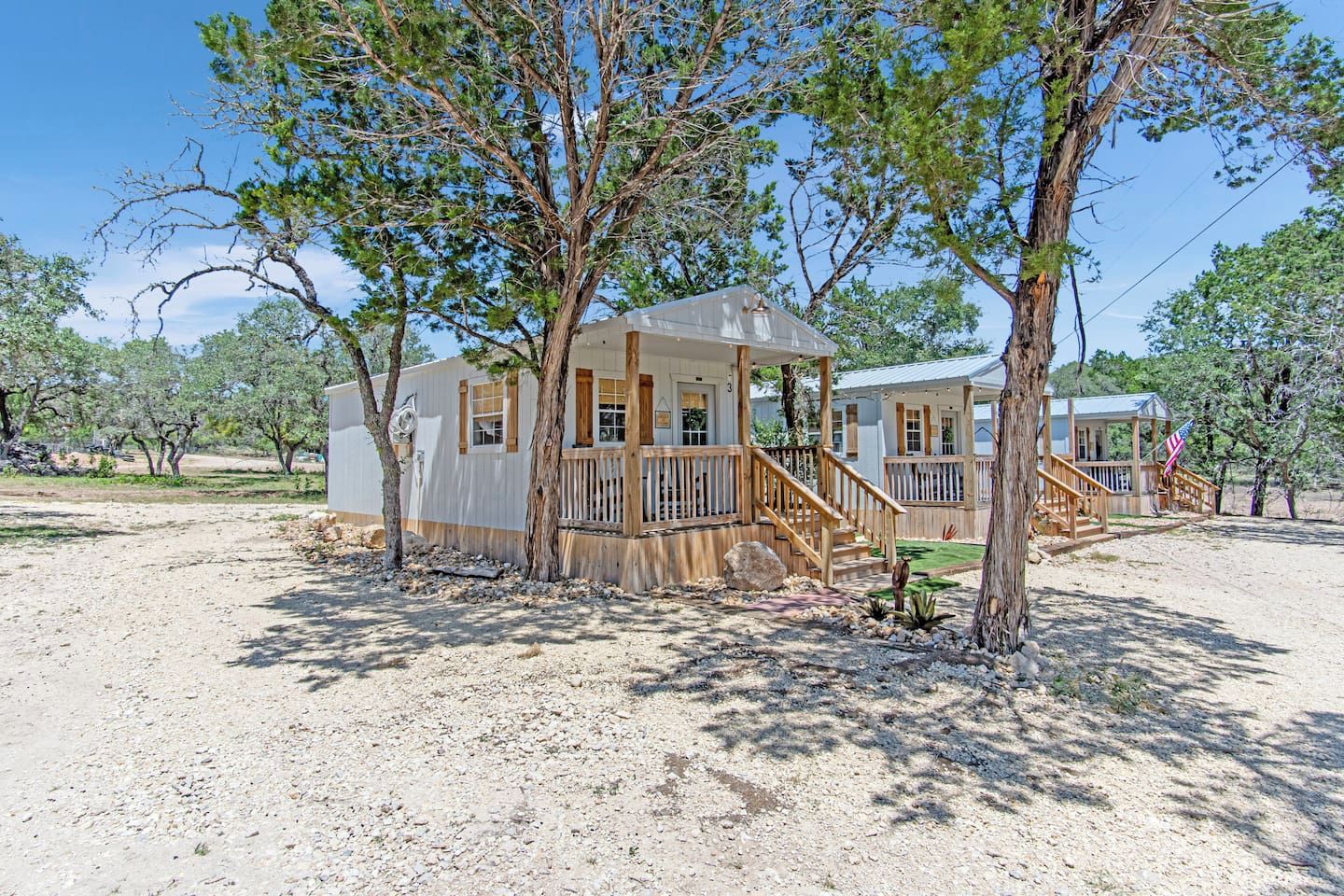 Uma casa móvel com varanda e um pequeno deck, rodeada por árvores e um chão de cascalho.