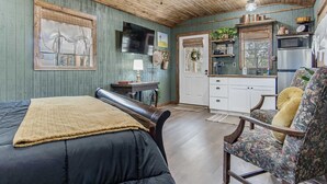 A cozy room with a bed, a chair, a desk, a television, a kitchen area, and a ceiling fan. - Lucky Star Ranch - Stargazer (Bandera)