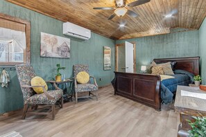 A room with a wooden ceiling, a bed with dark bedding, two floral-patterned chairs, a small table with a plant, and a wall-mounted air conditioner.