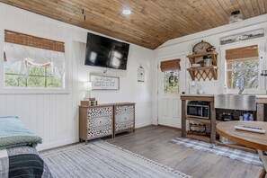 A cozy room with a wooden ceiling, a flat-screen TV mounted on the wall, a wooden cabinet with decorative cutouts, a round wooden table, and a window with a view of greenery. - Lucky Shooting Star #3 | Romantic Stay Near Parks (Bandera)