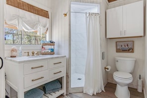 A bathroom with a white sink, vanity, and toilet. There is a shower curtain, a basket of toiletries, and a framed picture on the wall.