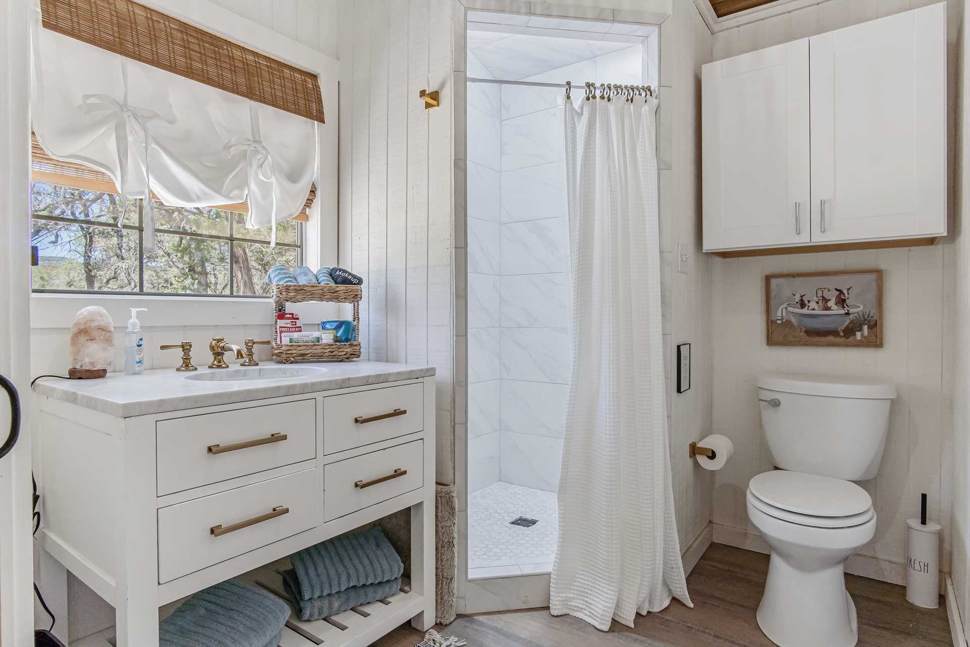 A bathroom with a white vanity, a shower with a white curtain, a toilet, and a window with a view of trees.