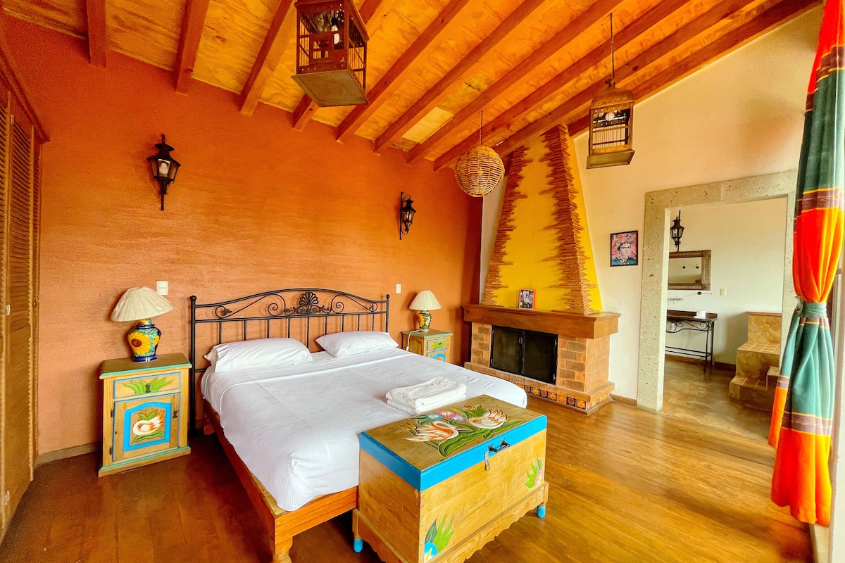 A bedroom with a wooden bed, a nightstand with a lamp, a dresser, a fireplace, and a wooden ceiling.