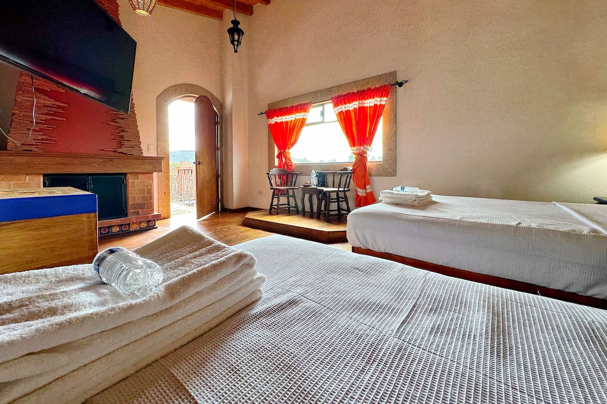 A room with two beds, a fireplace, a dining area with a table and chairs, and a view of the outside.