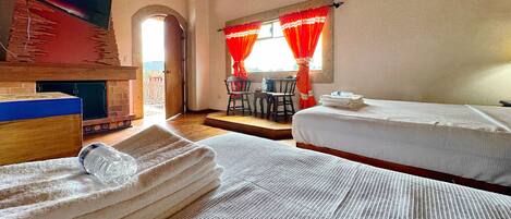 A room with two beds, a fireplace, a dining area with a table and chairs, and a view of the outside.