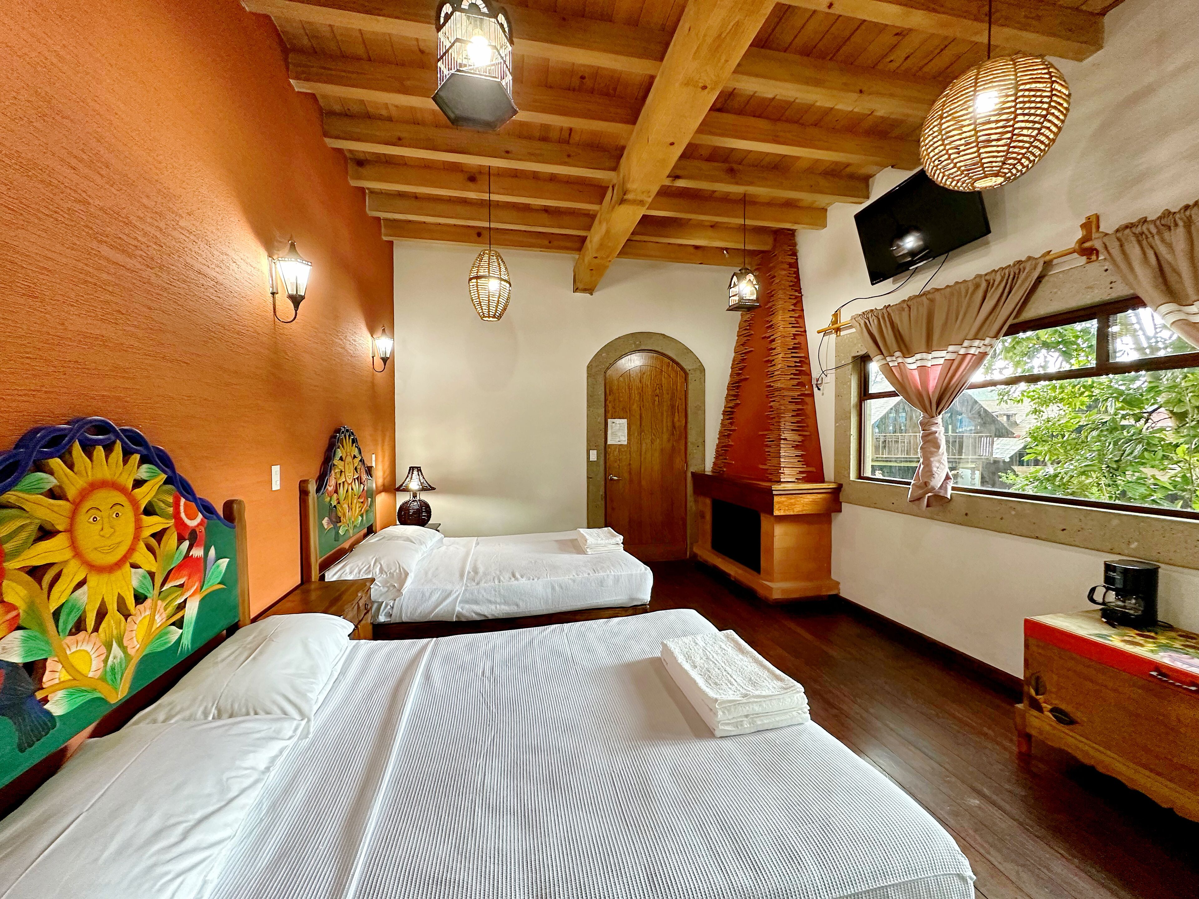 A room with two beds, a wooden ceiling, a fireplace, a TV, and a window with curtains.
