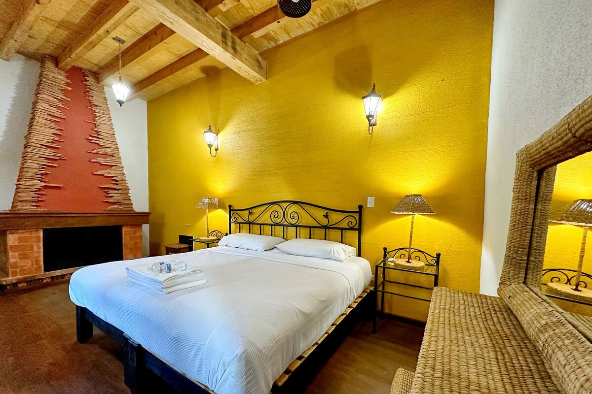 A bedroom with a bed, two bedside lamps, a small table, a chair, and a fireplace.