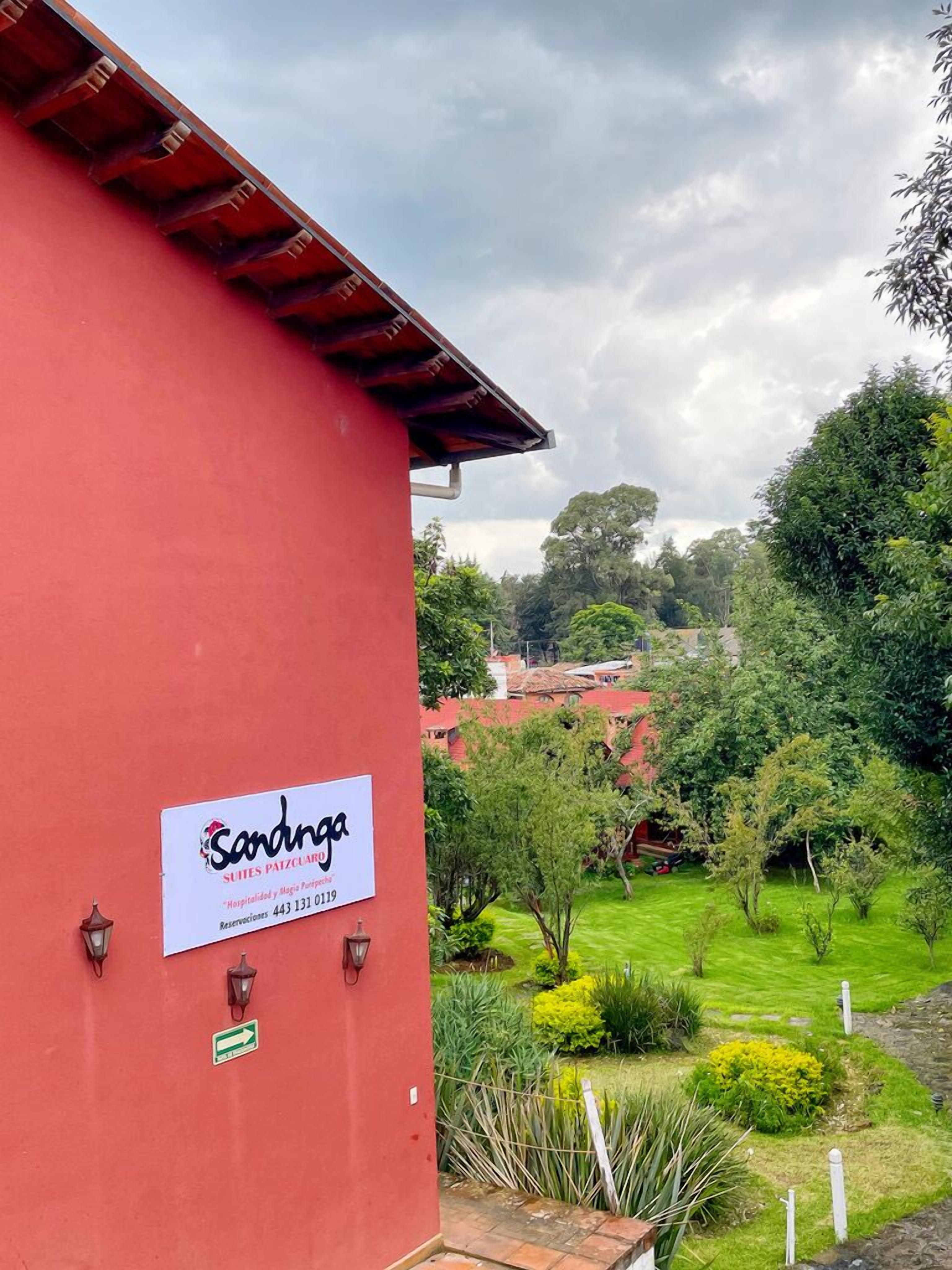 A red building with a sign for 'Sandinga Suites Patezcuaro' and a well-maintained garden with various plants and a pathway.