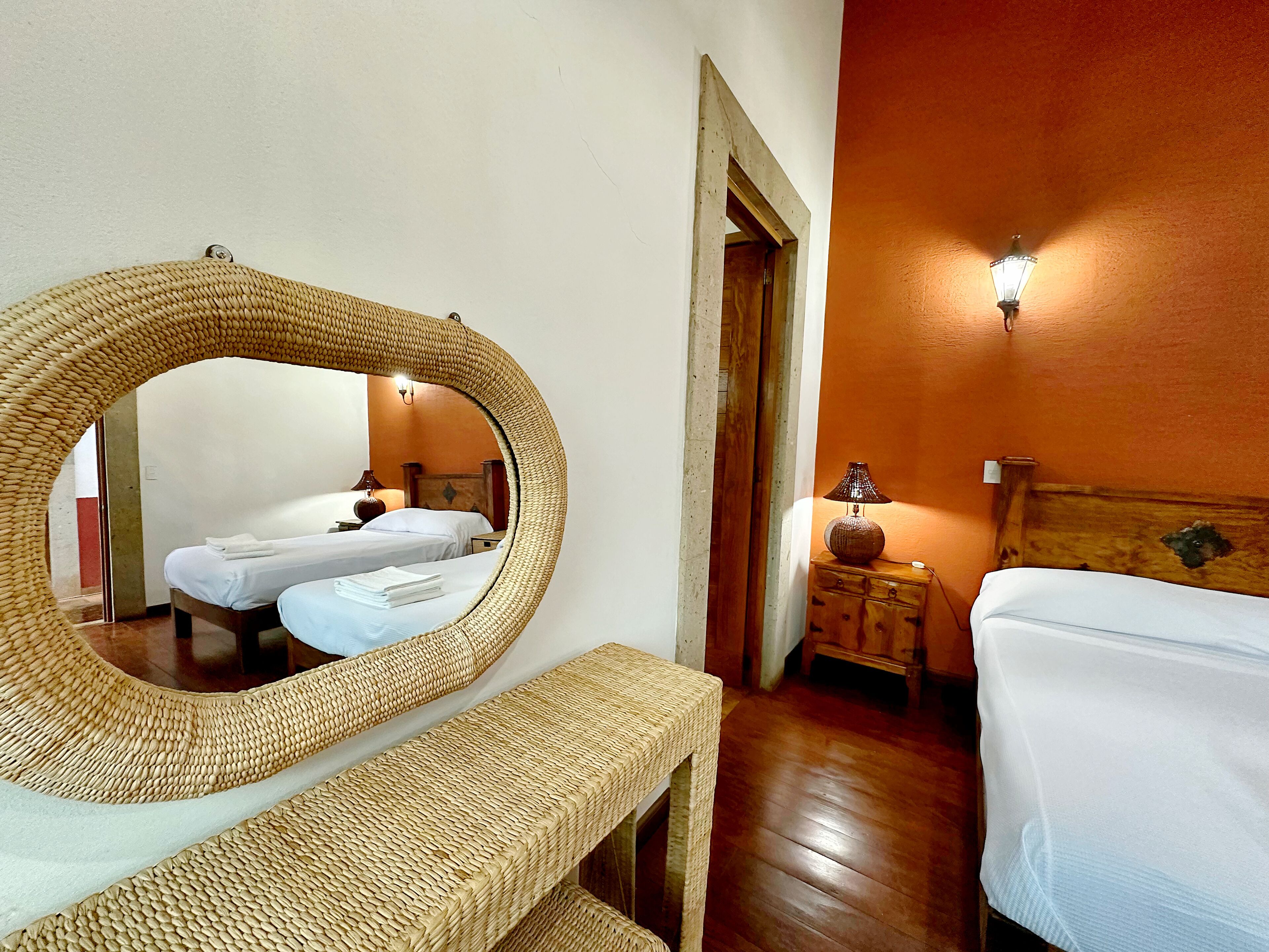 A room with two beds, a wicker bench, a wooden nightstand, and a wall-mounted lamp.
