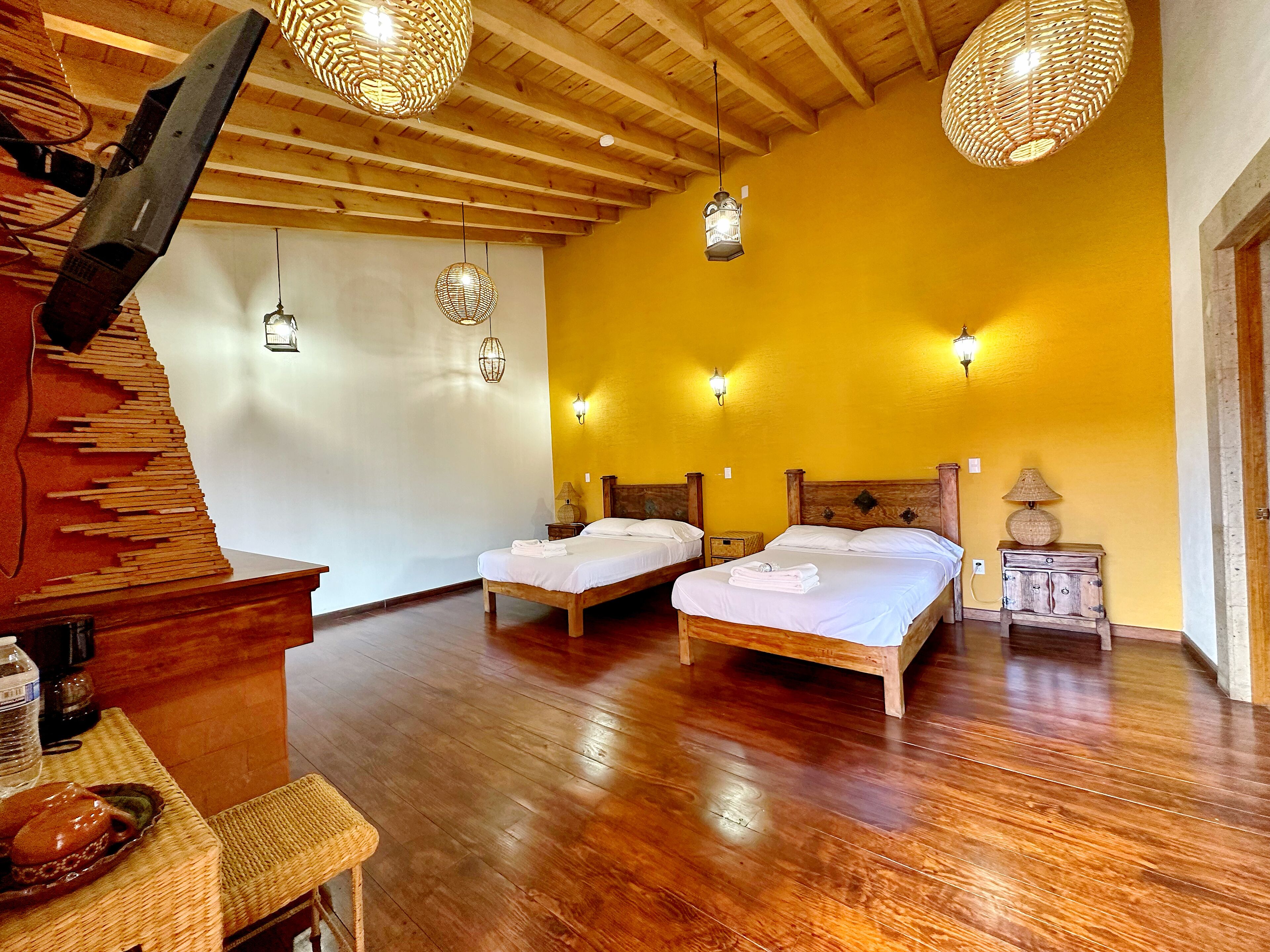 Habitación de hotel con piso de madera, dos camas, un aparador de madera, un televisor y luces fijadas a la pared.