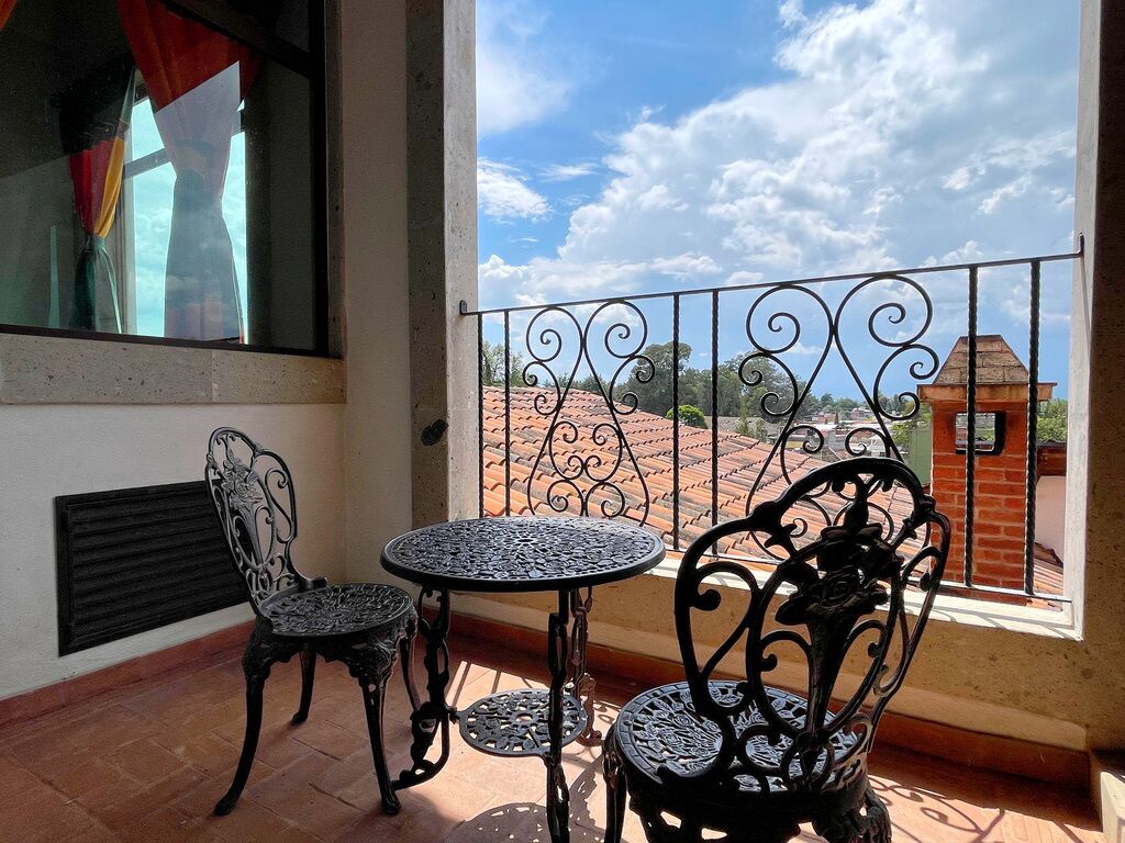 A balcony with two ornate metal chairs and a small round table, a window with curtains, and a view of a red-roofed building.