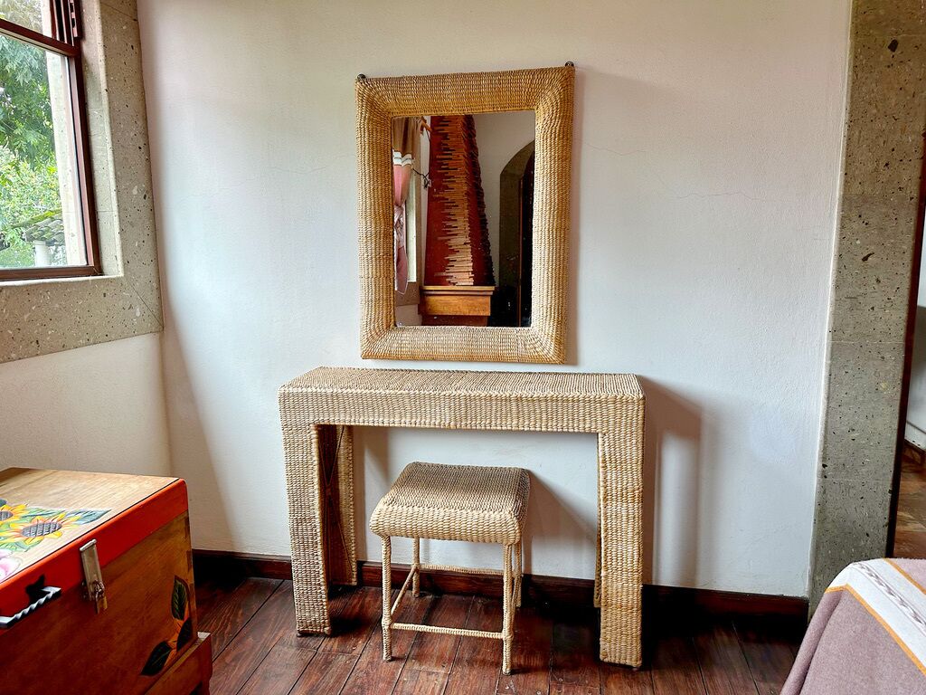 A room with a woven console table, a stool, and a mirror.