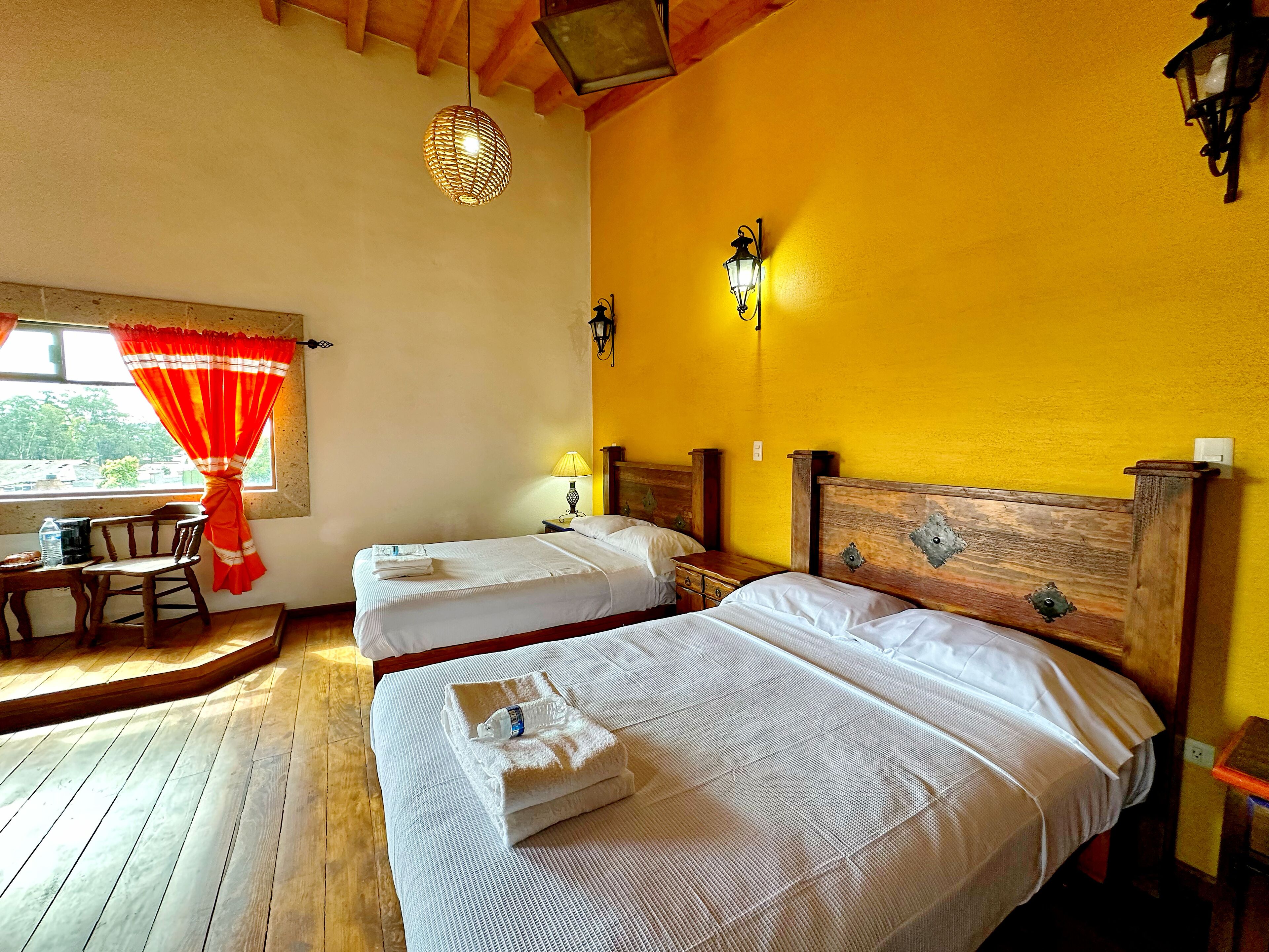 A room with two beds, a wooden floor, a window with red curtains, and wall-mounted lights.
