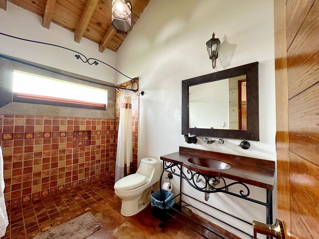Un baño con piso de baldosas, inodoro blanco, tocador de madera con espejo y lámpara colgante.