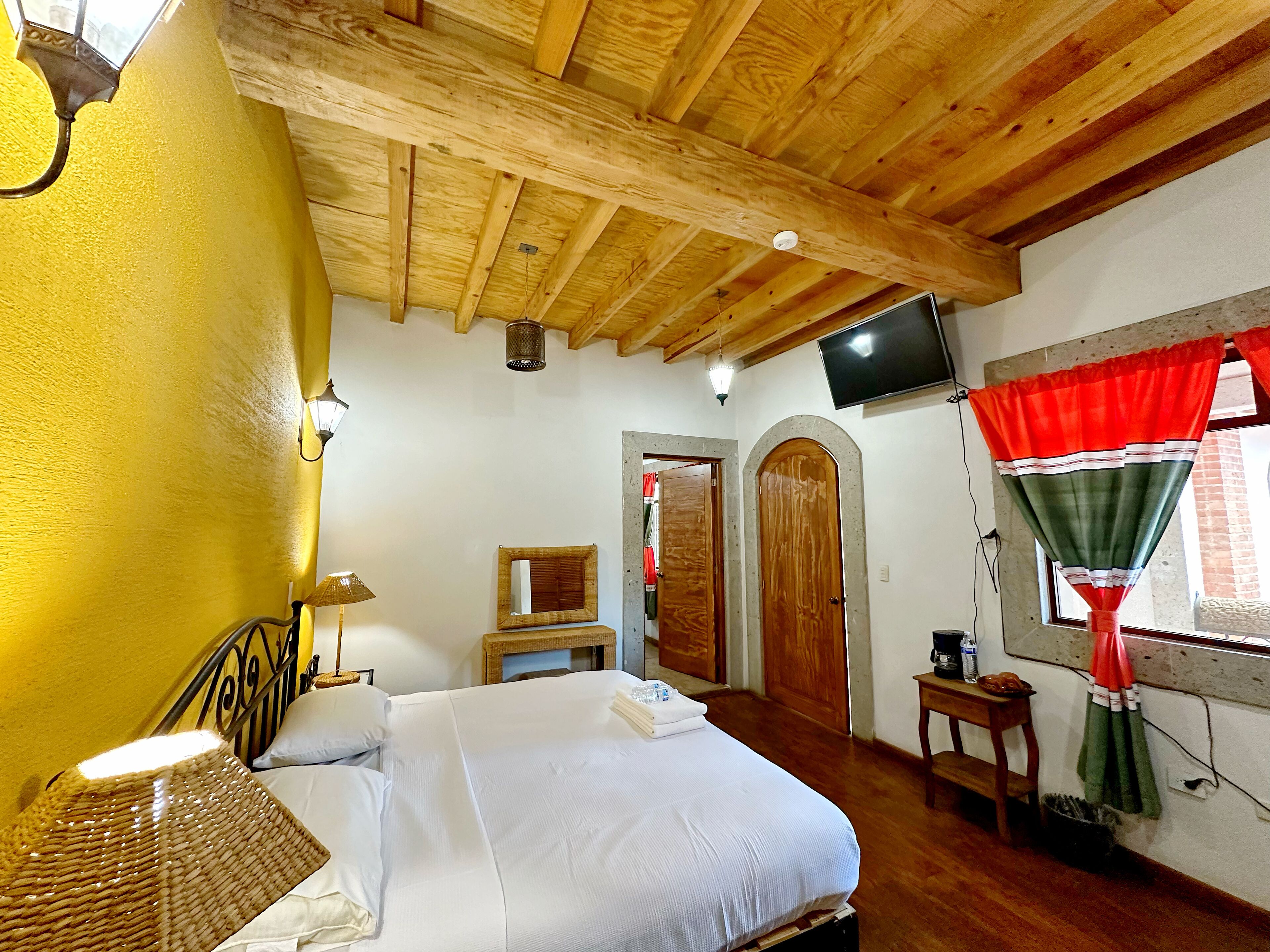 Une chambre avec un plafond en bois, un lit avec une literie blanche, une table de chevet, une lampe, un miroir et une fenêtre avec des rideaux.