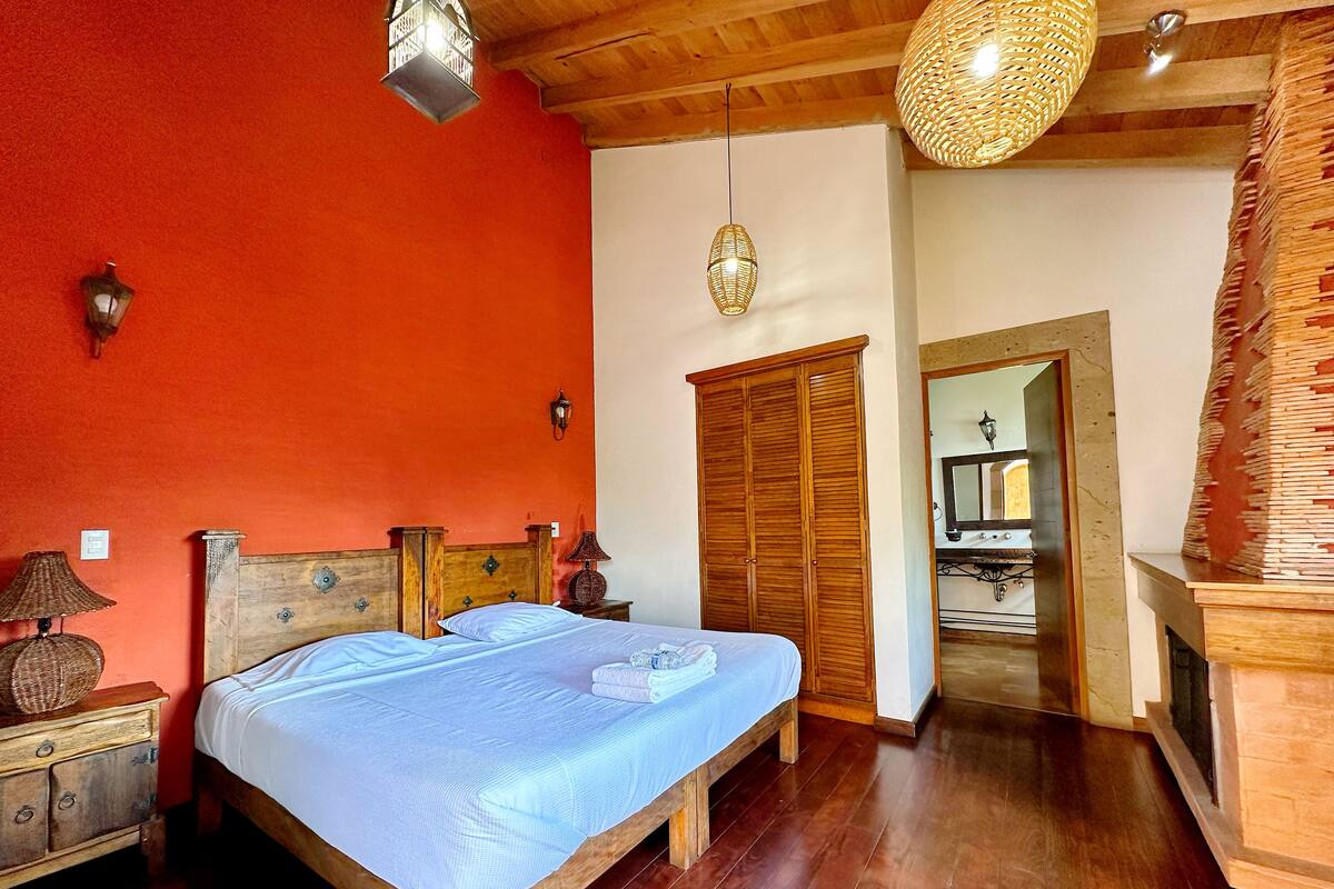 A bedroom with a wooden bed, a wardrobe, a nightstand with a lamp, and a hanging light fixture.
