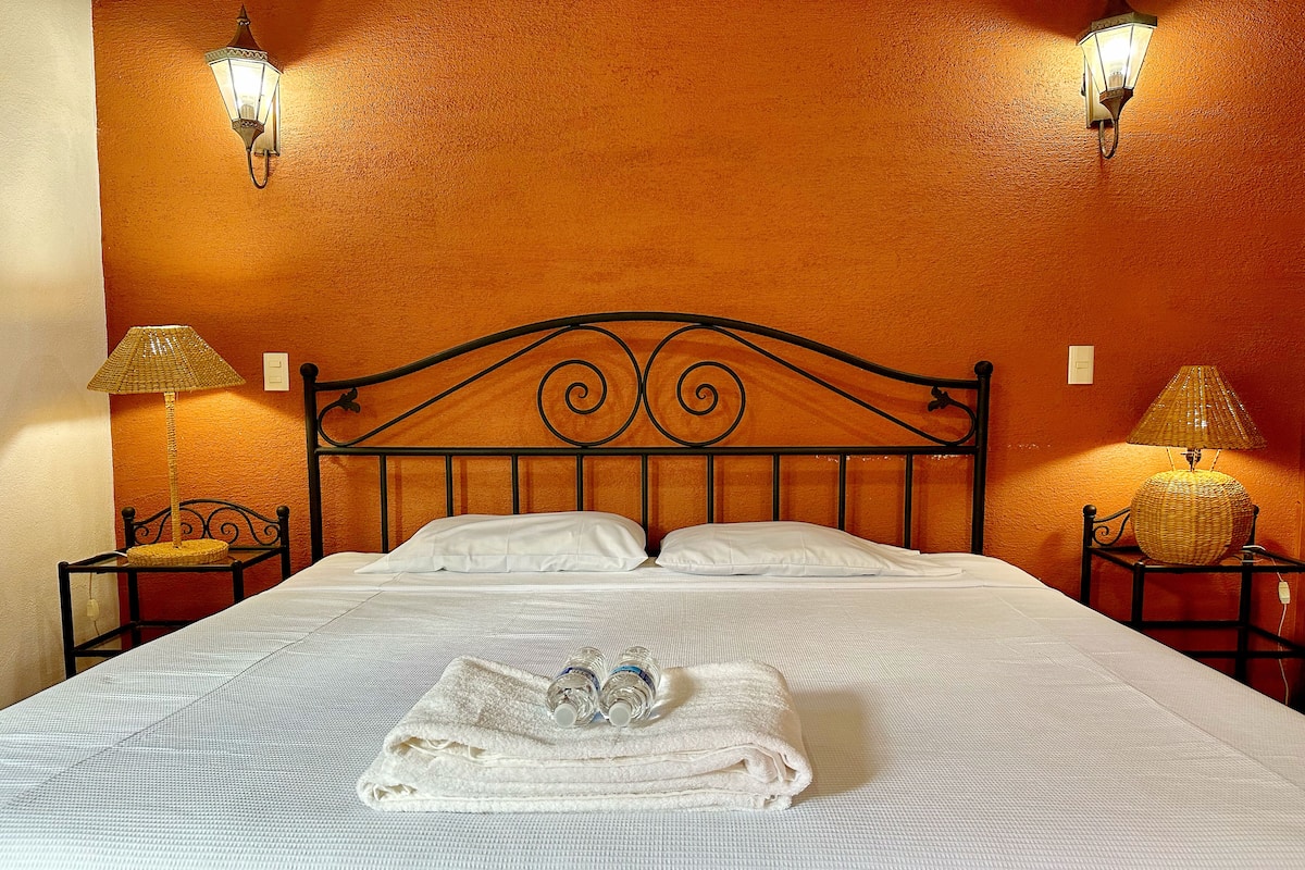 A neatly made bed with white linens, a folded towel, and two bottles of water on the bed. There are two nightstands with lamps and a wall sconce.