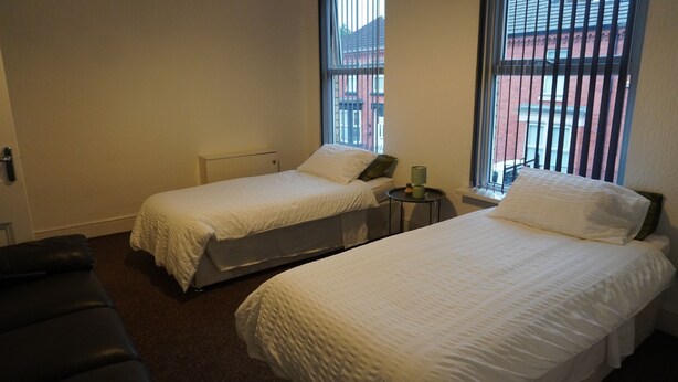 A room with two single beds, a small round table with a lamp, and a view of buildings through large windows.