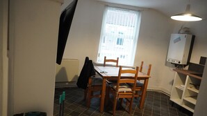 A small, compact room with a dining table and chairs, a radiator, a window with blinds, a white cabinet, and a ceiling light fixture.