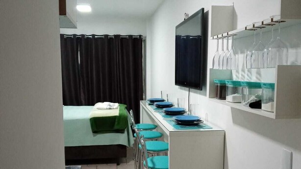 A compact hotel room with a bed, a small kitchenette, a dining area with chairs, a TV, and a shelf with glasses. - Apar Aconchegante no Centro de Curitiba  (Curitiba)