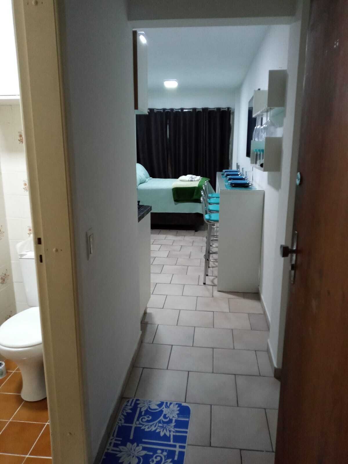 A narrow hallway leading to a bedroom with a bed, a desk with chairs, and a bathroom with a toilet.